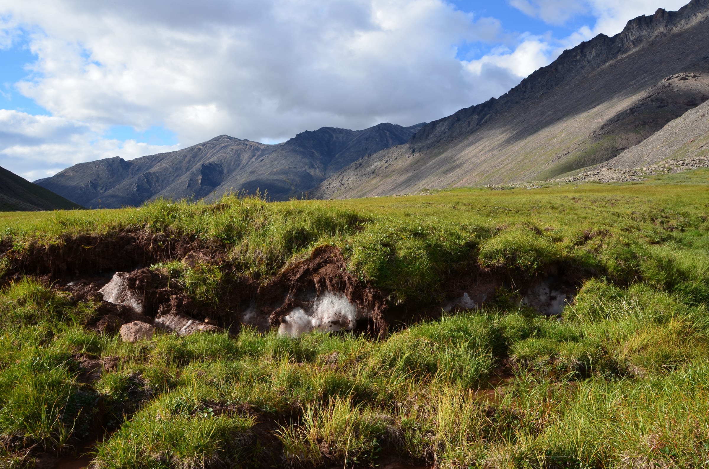 Exposed ground ice beneath tundra grass, with a mountain valley and sky beyond.